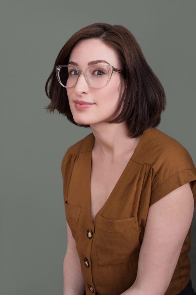 This is a theatrical headshot for Meredith. She is playing a bookish, smart character. Her smooth dar brown hair reaches just above her shoulders and is curled in. She's wearing clear plastic glasses with a large frame. Her lips are a pale wine color and her eyebrows are nicely shaped. She's facing to he left and her gaze is at the camera. She has a slight, pleasant smile. Her shirt is light brown and unremarkable. This is clearly someone who cares more about pursuits of the mind than her wardrobe. Photo taken by Rebecca Little Photography Pasadena, CA.